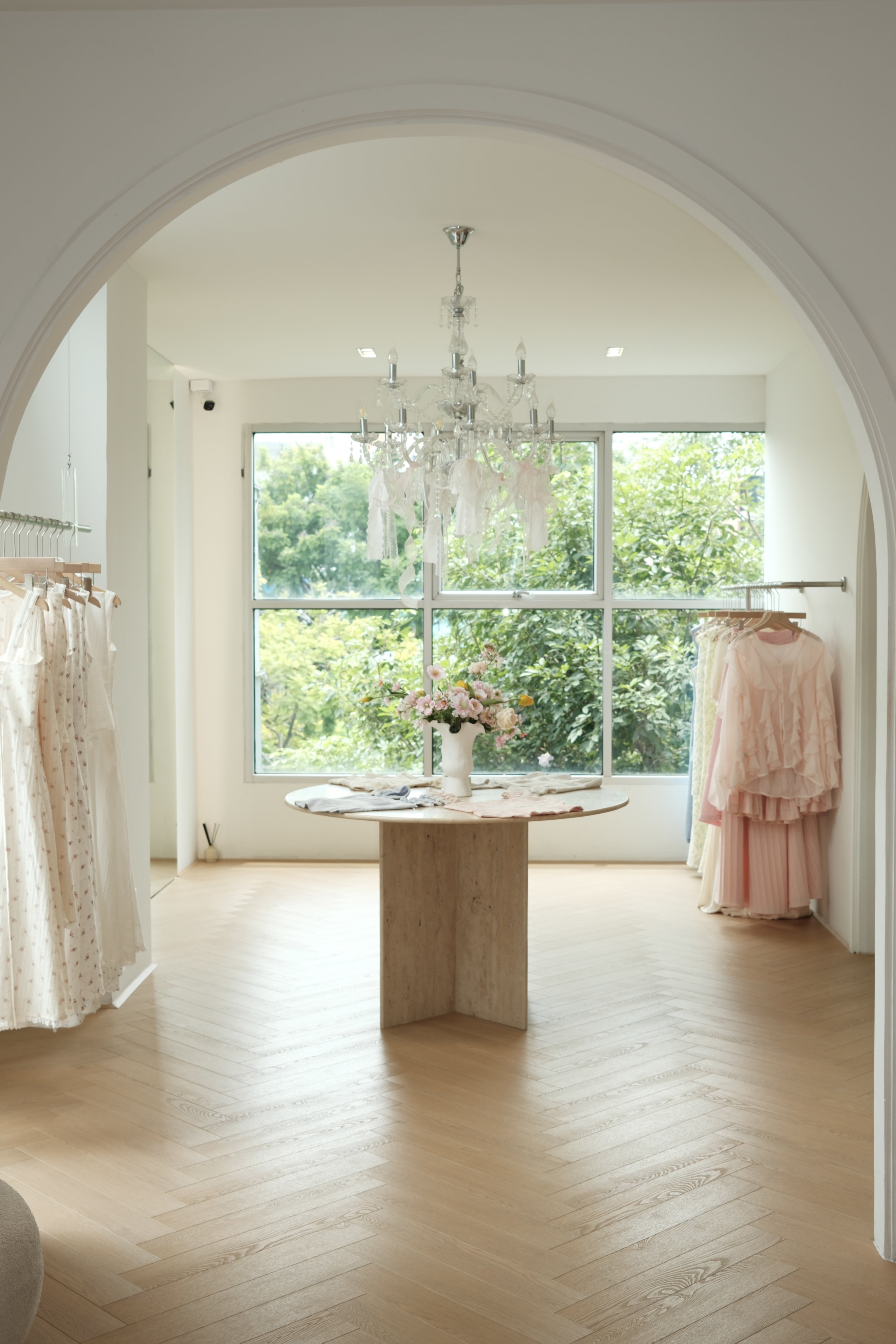 View through an arched doorway into the RAVii boutique, with a travertine table, crystal chandelier and rails of pink and cream dresses